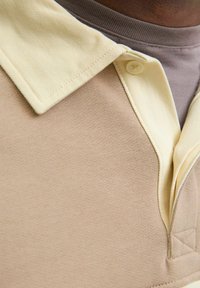 Polo majica s bež tijelom i svijetložutim rebrastim ovratnikom. Ima dvostruko dugme na prednjem dijelu i teksturiranu tkaninu za udobnost.