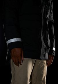 Black insulated jacket with a zipper, featuring a silver reflective strip on the sleeve. Paired with beige shorts, showcasing a casual look.