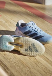 Paar hellblaue Adidas-Sneaker auf einem Holzboden, ein Schuh zeigt die sechseckig gemusterte Sohle mit sichtbarem Adidas-Logo.
