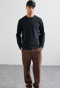 Sort langærmet skjorte med diskret logo, parret med brune plissee-bukser. Model iført en sort baseballcap, stående foran en neutral baggrund.