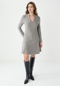 Grijs langemouwen jurkje met een V-hals, voorzien van gestructureerde naden en een A-lijn silhouet. Gepaard met zwarte kniehoge laarzen met hak.