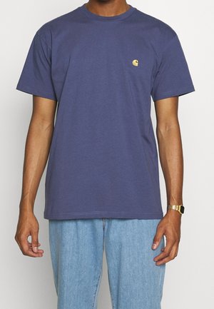 T-shirt en coton bleu marine à manches courtes, coupe décontractée, avec petit logo doré sur le côté gauche de la poitrine. Porté avec un jean en denim bleu clair.