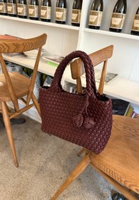 Borsa di stoffa bordeaux con manici arrotondati, caratterizzata da accenti annodati, posata su una sedia di legno con finitura rustica.