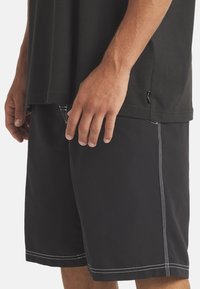 Mustat uimaportit valkoisella ompelutavalla, sileä pinta ja sivutaskut, yhdistettynä löysään mustaan t-paitaan.