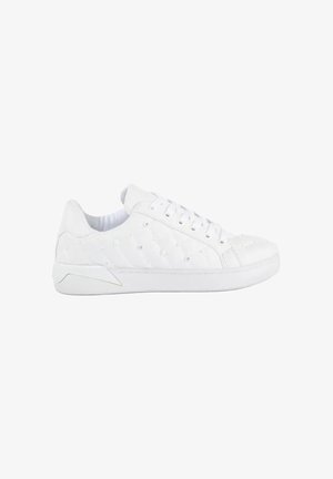 Witte lage sneaker met doorgestikt patroon, vetersluiting en een platte rubberen zool, gezien van opzij op een witte achtergrond.