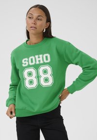 Zelená mikina s potiskem textu "SOHO" a číslic "88" v bílé barvě. Má kulatý výstřih a žebrované manžety. Nosená s černými kalhotami.