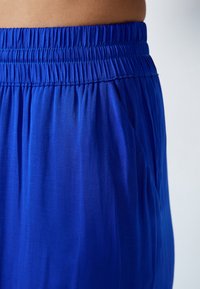 Pantalons légers bleu avec une taille élastique et des poches latérales. Le tissu a une texture lisse avec un léger brillant.