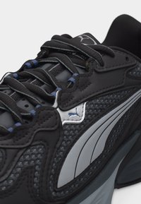 Κοντινό πλάνο μαύρου και γκρι αθλητικού παπουτσιού Puma με υφασμένο πλέγμα, χοντρές κορδόνια και λογότυπο στο πλάι και στη γλώσσα.