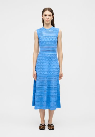 Femme debout, portant une robe midi sans manches à motif bleu et des ballerines à imprimé léopard, sur un fond blanc uni.
