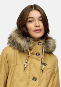 Żółty zimowy płaszcz z kapturem z sztucznego futra. Posiada drewniane i metalowe akcenty, zapięcia na guziki oraz gładką fakturę. Kołnierz jest puszysty i ciepły.