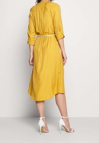 Żółta sukienka do kolan z rękawami o długości trzech ćwierci i marszczonym tyłem. Posiada wcięcie w talii z białym pasem oraz asymetryczny dół.