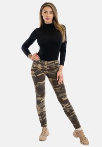 Body a maniche lunghe nere abbinato a jeans a vita alta con motivo camouflage e due bottoni. La modella indossa scarpe beige.