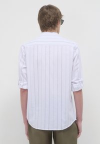 Mann mit kurzen braunen Haaren, der eine Sonnenbrille trägt, ein weißes Langarmshirt mit feinen vertikalen Streifen und olivgrüne Hose an hat, von hinten blickt.