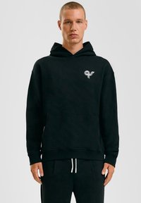 Zwarte capuchontrui gemaakt van zachte stof, met een voorzak en een klein grafisch logo op de linkerborst. Textured afwerking.