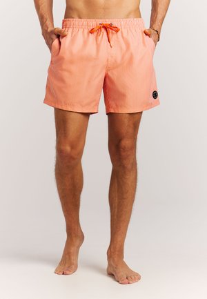 Man die mouwloze oranje-wit gestreepte zwembroek met elastische tailleband en voorzakken draagt, staat blootsvoets tegen een effen achtergrond.