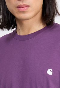 Paarse katoenen T-shirt met een ronde hals en korte mouwen; heeft een klein wit logo op de linkerborst. Zachte textuur.