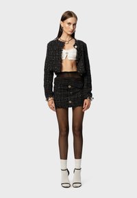 Giacca cropped in tweed nero, top corto bianco testurizzato, mini gonna nera con bottoni, collant neri trasparenti e calzini alla caviglia bianchi aperti con dettagli.