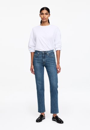 Kobieta stojąca przodem, ubrana w białą koszulę z długim rękawem, niebieskie dżinsy z wysokim stanem o prostym kroju oraz czarne skórzane mokasyny na białym tle.