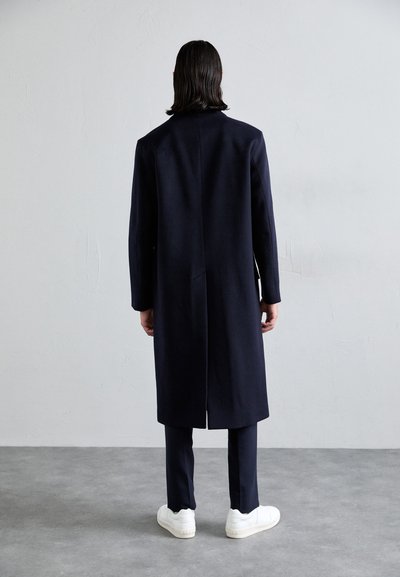 Cappotto di lana navy con taglio dritto, maniche lunghe, dettaglio di apertura sul retro, abbinato a pantaloni scuri e sneakers bianche su un pavimento grigio.