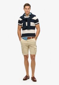 Camiseta polo a rayas en azul marino y blanco con mangas cortas y un suéter azul marino colgado sobre los hombros, pantalones cortos beige tipo cargo, mocasines marrones.