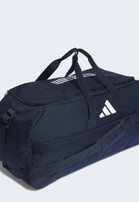 Námořnická modrá sportovní taška vyrobená z odolné tkaniny. Obsahuje bílé logo a pruhy, zipový uzávěr, dvojité uši a nastavitelné ramenní popruhy.