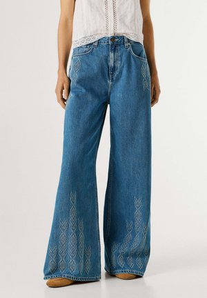 Vrouw draagt een hooggetailleerde, wijdlopende blauwe spijkerbroek met lichte geborduurde patronen op de dijen en zoom, gecombineerd met een wit mouwloos topje.