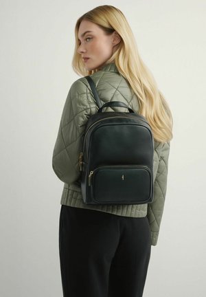 Blondynka w zielonej pikowanej kurtce i czarnych spodniach, niosąca na plecach czarny skórzany plecak ze złotymi suwakami.