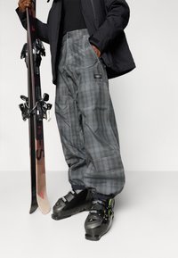 Oseba v sivih kariranih smučarskih hlačah, črnih smučarskih škornjih in črni jakni drži par smuči pokončno na belem ozadju.