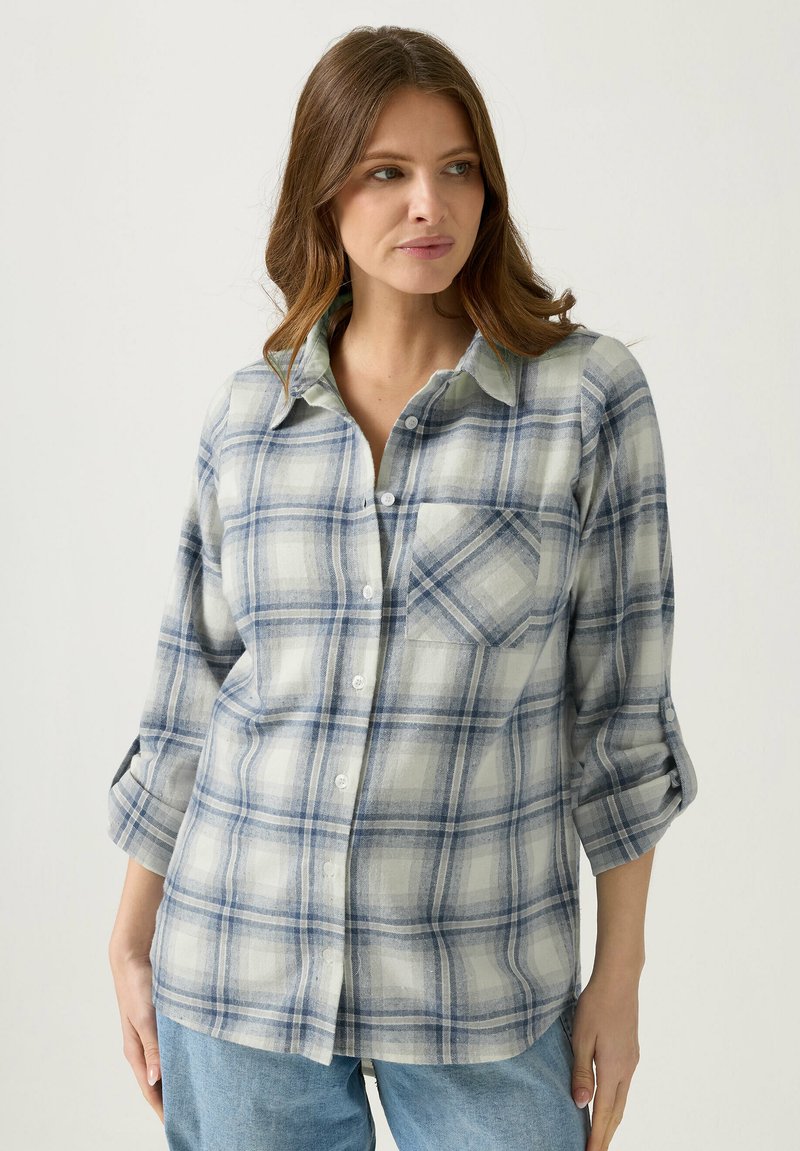 Nainen, jolla on sinivalkoruudullinen nappipaita, jonka hihat on käännetty ylös, ja vaaleansiniset farkut, katsoo sivulle.