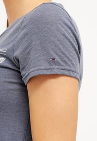 Camiseta de algodón gris con mangas cortas, que presenta un pequeño logo bordado en la manga. Textura suave, diseño casual, color sutil.