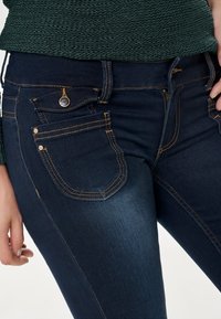 Jean skinny bleu foncé en denim avec des coutures dorées. Dispose de poches avant, d'une fermeture à bouton et d'une texture lisse, légèrement brillante.