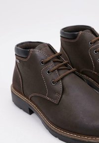 Botas de tobillo de cuero marrón con cuello acolchado, cordones marrones, ojales de metal, costuras detalladas y suela de goma gruesa.