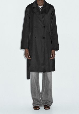 Personne portant un manteau long croisé noir sur un jean gris à jambes larges et des sandales noires à bouts ouverts, debout devant un fond uni.