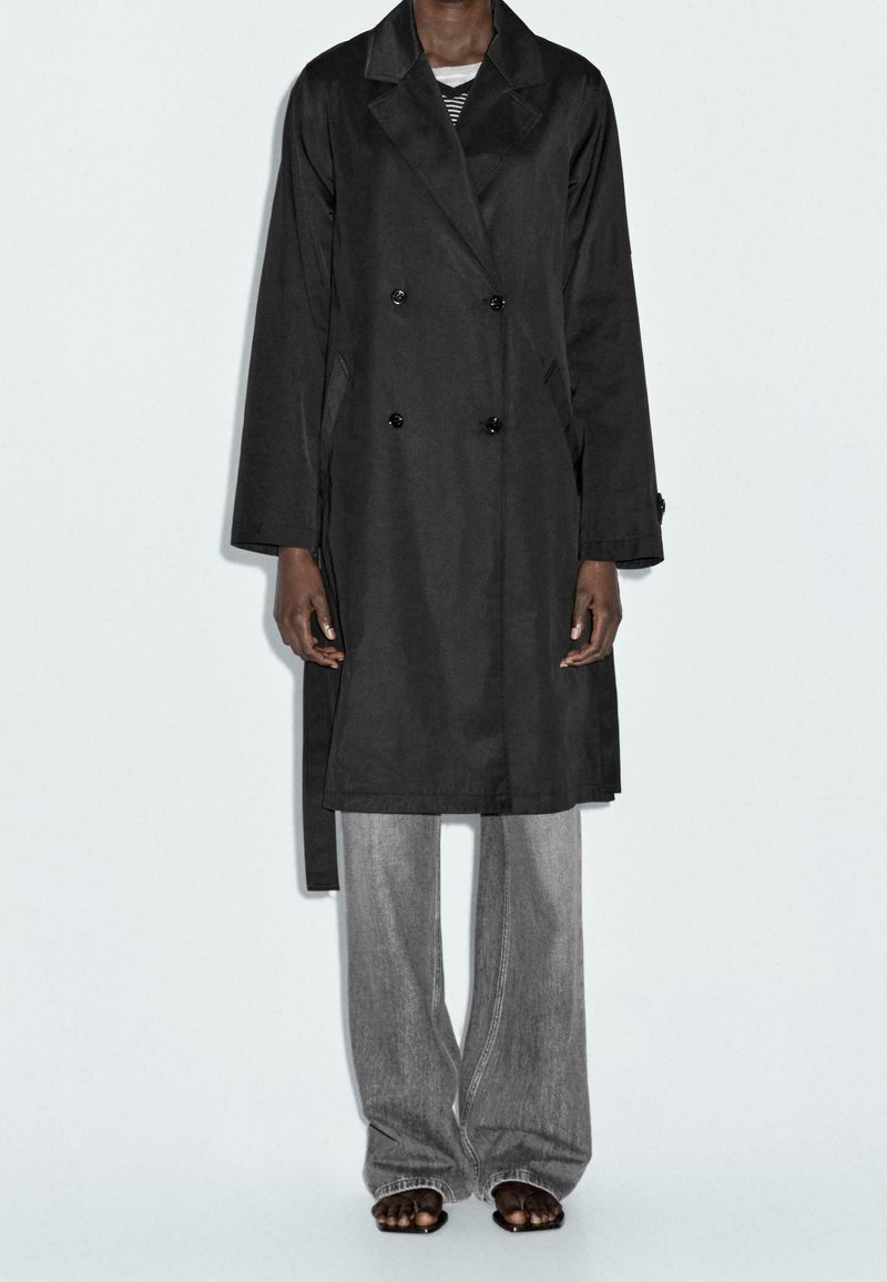 Personne portant un manteau long croisé noir sur un jean gris à jambes larges et des sandales noires à bouts ouverts, debout devant un fond uni.