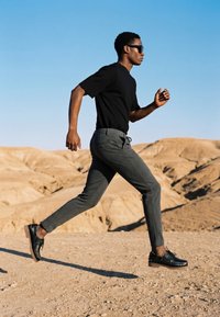 Homme portant des lunettes de soleil, une chemise noire et un pantalon gris courant sur un terrain désertique avec des collines sablonneuses et un ciel bleu clair en arrière-plan.