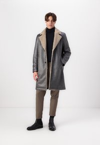 Abrigo gris con un acabado texturizado y cuello de borrego beige, diseño de un solo botón, combinado con pantalones marrones y botas negras.