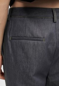 Οπίσθια όψη σκούρου γκρι τζιν παντελονιού με κίτρινη ραφή και μία μόνο τσέπη στο πίσω μέρος, στην περιοχή του ισχίου και της κάτω πλάτης ενός ατόμου.