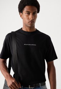 Black cotton t-shirt featuring a crew neck, short sleeves, and white "SCOTCH & SODA" logo on the front. Model wears a shoulder bag.