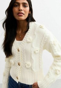 Cardigan lavorato a maglia crema con decorazioni floreali, design a trecce, polsini a costine e bottoni in legno. Abbinato a una maglia leggera e jeans blu.