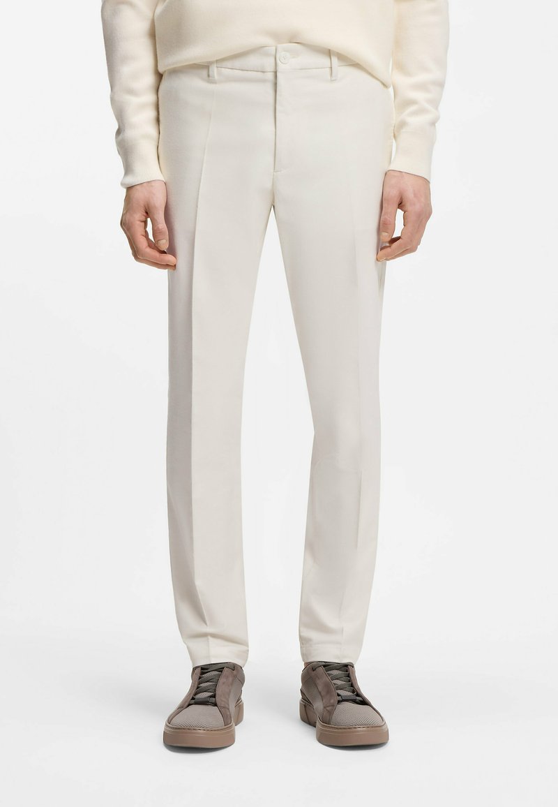 Άνδρας που φοράει παντελόνι σε εκρού χρώμα και δερμάτινα αθλητικά παπούτσια ταμπά με κορδόνια, στέκεται με τα χέρια χαλαρά δίπλα στο σώμα μπροστά σε λευκό φόντο.