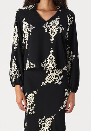Vrouw die een zwart V-hals top met lange mouwen en bijpassende rok draagt met crèmekleurige, sierlijke bloemenpatronen.