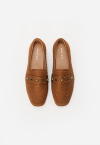 Mocassins en suede marron au design plat, ornés de clous décoratifs et d'un bandeau avec des accents noirs et dorés. Texture douce et lisse dans l'ensemble.