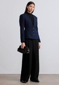 Μπλε ναυτικό σακάκι με υφή και βαθύ V-λαιμόκοψη, τρία χρυσά κουμπιά, συνδυασμένο με μαύρο παντελόνι φαρδιάς γραμμής και μικρή μαύρη υφασμάτινη τσάντα ώμου.