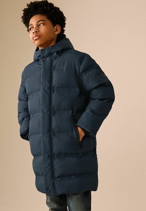 Teen wearing a dark blue padded winter coat with hood, standing with hands in pockets, looking to the side against beige background.