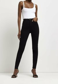 Jeans skinny negros de cintura alta con una textura de denim suave; cuenta con un cierre frontal con botón y presillas para cinturón. Combinados con una camiseta sin mangas blanca de canalé.