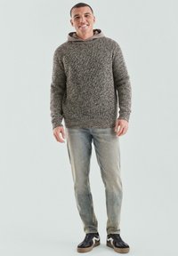 Sweter dzianinowy w brązowo-beżowych odcieniach z kapturem, zestawiony z jasnoniebieskimi dżinsami i czarnymi trampkami z brązowymi akcentami. Model stoi.