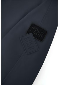 Marinfärgad tyg med två fyrkantiga "POLO CLUB" lappar, en upphöjd i svart gummi och en i strukturerad svart tyg. Slät yta med sydd söm.