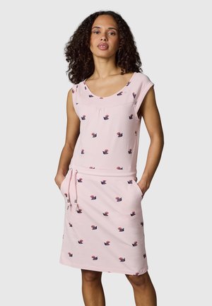 Kobieta o kręconych włosach, ubrana w jasnoróżową sukienkę bez rękawów z drobnymi kwiatowymi wzorami, z rękami w kieszeniach na jednolitym tle.