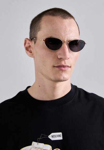 Round, black sunglasses with thin gold metal frames. The lenses are dark and reflective. Black t-shirt with a graphic and logo visible.