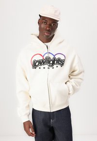 Kremo spalvos zip-up maikutė su dideliu juodu piešiniu, kurį papildo raudonos, violetinės ir mėlynos akcentai, derinama su tamsiais džinsais. Nešiojama šviesios spalvos kepurė.